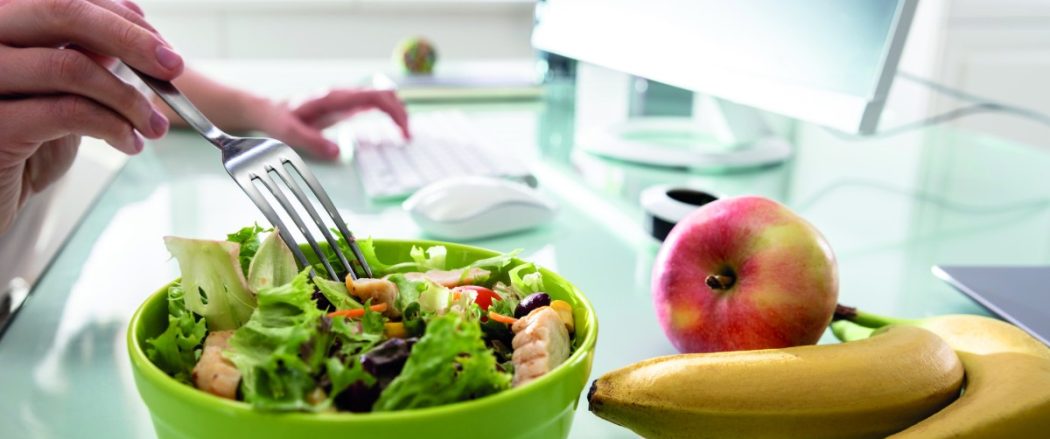 This image shows a healthy workplace lunch setting. A person is eating a fresh salad with lettuce, grilled chicken, and vegetables while working on a computer. On the desk, there are also bananas and an apple, emphasizing a nutritious diet.