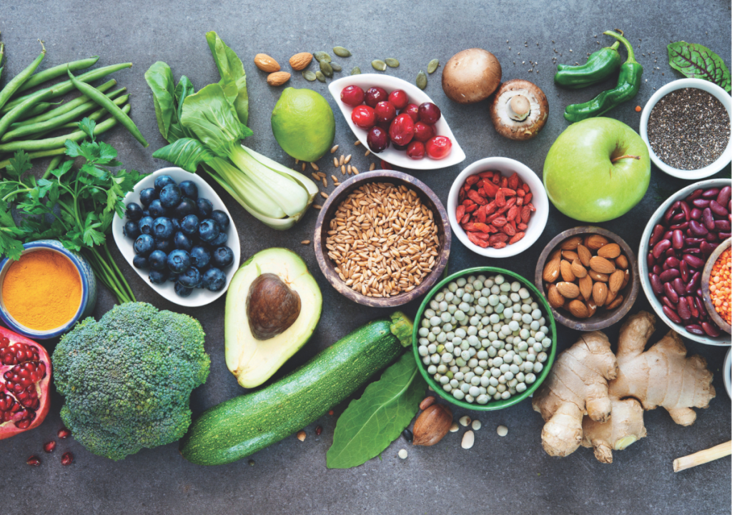 This image showcases a variety of fresh, healthy foods, including fruits, vegetables, legumes, nuts, seeds, and whole grains. The arrangement features ingredients like blueberries, avocado, beans, lentils, almonds, broccoli, and turmeric, emphasizing a colourful and nutrient-rich diet.