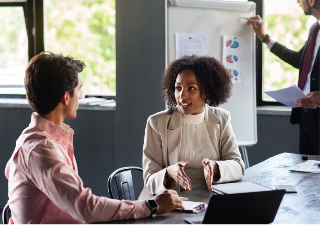 This image shows a professional business meeting in an office setting. A woman in a beige blazer is engaged in conversation with a male colleague, using hand gestures while speaking.